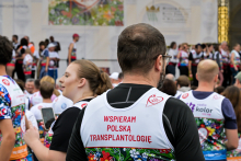 Mężczyzna w okularach stoi tyłem do kamery. Nosi białą koszulkę z czerwonym napisem „WSPIERAM POLSKĄ TRANSPLANTOLOGIĘ”. W tle widać kilkanaście osób w kolorowych strojach sportowych. Za nimi znajduje się duży baner z grafiką i nieczytelnymi napisami. Scena odbywa się na zewnątrz.
