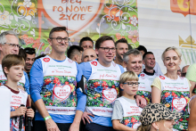 Około dwudziestu osób pozuje na tle kolorowego baneru z napisem „Bieg po nowe życie”. Mężczyzna w niebieskiej koszulce z numerem 48 stoi obok chłopca w białej koszulce. Uczestnicy noszą koszulki z sercem i hasłem „Razem dla transplantacji”. W tle widać dorosłych i dzieci w podobnych strojach.