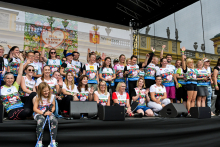 About thirty people are standing on a covered stage. They are wearing colorful T-shirts with the logo “Run for a New Life.” They are smiling and waving at the camera. Behind the stage, there is a building with sculptures on the roof.