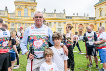 Mężczyzna w średnim wieku trzyma kijek do nordic walking i trzyma lewą rękę na ramieniu chłopca. Oboje stoją na trawie przed żółtym budynkiem z ozdobnymi elementami. Mężczyzna nosi koszulkę z napisem „Bieg po nowe życie”. W tle widać kilkanaście osób w koszulkach z napisem „Polska Transplantologia”. Niebo jest pochmurne.
