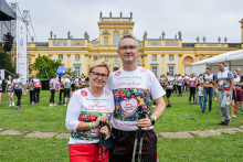 A woman and a man wearing white T-shirts with colorful prints are standing on the grass with Nordic walking poles. In the background, you can see a yellow palace with decorative windows. Behind them stands a group of people wearing similar sports outfits. 