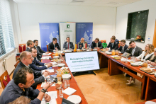 Kilka osób siedzi wokół dużego prostokątnego stołu w sali konferencyjnej. Wszyscy są elegancko ubrani. Na stole leżą dokumenty i laptopy. Na końcu stołu widać duży ekran z napisem „Reimagining University International Action – Territorial Diplomacy and Decentralized Cooperation from the Global South”. W tle znajdują się dwa banery z częściowo widocznymi logo i tekstem.