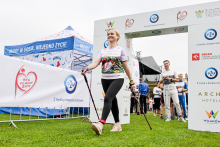 Kobieta w białej koszulce z kolorowym nadrukiem i czarnych legginsach przechodzi przez bramkę z logotypami sponsorów. Trzyma kijki do nordic walking. W tle widać namiot z napisem „Mamy w sobie, niejedno życie” oraz baner z hasłem „Bieg po nowe życie”. Trawa jest zielona, niebo pochmurne.