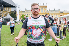 Mężczyzna w kolorowej koszulce z napisem „Bieg po nowe życie” i logo „Polska Transplantologia” stoi na trawie przed pałacem. Na koszulce widnieje napis „Krzysztof Zojka”. Mężczyzna trzyma kijki do nordic walking. W tle widać uczestników wydarzenia, namioty i scenę. Pogoda jest pochmurna.
