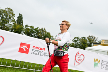 Kobieta w średnim wieku idzie z kijkami do nordic walking. Ma krótkie blond włosy, okulary, białą bluzkę z kolorowym wzorem i czerwone spodnie. W tle widać baner z logotypem „ORLEN Fundacja im. Łukasiewicza” i napisem „Bieg po nowe życie”. Po lewej rosną drzewa, po prawej stoi namiot i budynek muzeum w Wilanowie. Nad sceną lata dron.