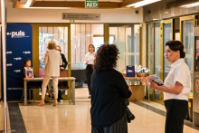Two women talking in a university hallway. The hallway is bright and well-lit. Other people can be seen in the background, some of them sitting at a table. Next to them is a blue wall with white lettering and the logo of the Regional Medical Chamber.