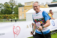 A middle-aged man is walking with Nordic walking poles. He is wearing a blue T-shirt with a colorful print and the words “Run for a New Life.” Next to him is a person wearing a yellow T-shirt. In the background, you can see a yellow building, a white tent, and a banner with a red heart symbol and the words “Run for a New Life.” .