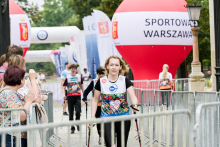 Kobieta z kijkami do nordic walking przekracza linię mety podczas zawodów w Warszawie. W tle stoi duży czerwono-biały balon z napisem „SPORTOWA WARSZAWA”. Po lewej stronie grupa osób bije brawo. Dalej widać uczestników biegu oraz bramę mety z logotypami sponsorów.