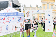 Mężczyzna trzyma za ręce dwoje dzieci. Wszyscy noszą koszulki z kolorowym nadrukiem i napisem „Bieg po nowe życie”. Stoją na zielonej trawie przed żółtym pałacem. W tle widać biały namiot z logo PZU oraz bramę startową z logotypami sponsorów. Pogoda jest pochmurna.