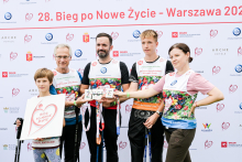 Pięć osób pozuje na tle ścianki z logo sponsorów podczas 28. Biegu po Nowe Życie w Warszawie. Trzy kobiety i dwóch mężczyzn noszą kolorowe koszulki sportowe i trzymają kijki do nordic walking. Czworo z nich trzyma tabliczki z napisem „Życie”, jedna kobieta trzyma plakat z sercem i napisem „Bieg po Nowe Życie”.