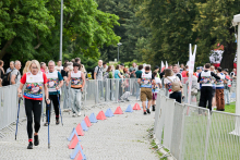 Grupa kobiet i mężczyzn idzie trasą nordic walking na świeżym powietrzu. Uczestnicy noszą kolorowe koszulki z numerami startowymi. Niektórzy trzymają kijki. Trasa jest oznaczona pachołkami i otoczona barierkami. W tle widać drzewa, widzów i innych uczestników. Jest słonecznie.