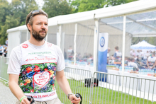 Mężczyzna uczestniczy w wydarzeniu sportowym. Nosi kolorową koszulkę z napisami „Bieg po nowe życie” i „Polska Transplantologia”. Trzyma kijki do nordic walking. Ma okulary przeciwsłoneczne na głowie i zegarek na lewym nadgarstku. W tle stoi biały namiot z przezroczystymi ścianami. W środku przebywają inni uczestnicy.