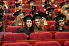 Graduates in gowns and mortarboards are seated in an auditorium; in the foreground, two people are smiling at the camera.