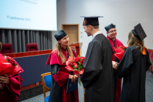 Two graduates in gowns present flowers to two individuals dressed in red robes.