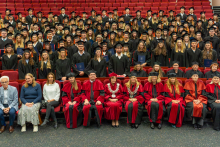A group of individuals in gowns and mortarboards is seated in an auditorium during the graduation ceremony; in the front row, people in academic attire, with graduates holding diplomas seated behind them.