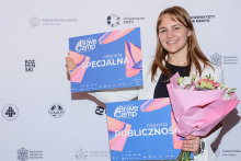 Magdala Halska. Młoda kobieta z długimi włosami stoi na tle ścianki z logotypami uczelni. Uśmiecha się. Nosi biały żakiet. Trzyma bukiet w różowym papierze i dwa plakaty z napisami „Brave Camp nagroda SPECJALNA” oraz „Brave Camp nagroda PUBLICZNOŚCI”.