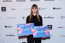 Kaja Tyszkiewicz. Młoda kobieta z długimi, prostymi włosami stoi na tle ścianki z logotypami BraveCamp, Politechniki Warszawskiej i Uniwersytetu SWPS. Nosi czarne ubranie. Trzyma dwa niebieskie plakaty z napisami „nagroda UWRC” i „nagroda SPECJALNA”.