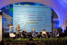 Pięć osób na scenie. To uczestnicy sesji tematycznej. Cztery osoby siedzą, jedna stoi i trzyma w rękach mikrofon. Za nimi widać ekran multimedialny.