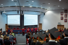 Six people in gowns are on stage during the diploma awarding ceremony; in front of them, graduates in caps are standing and taking the oath.