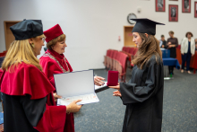 The female graduate in a gown receives a diploma and a medal from two individuals dressed in red robes.