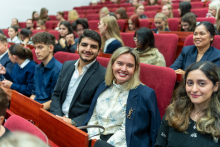 Four people are sitting in red armchairs in a lecture hall. Two women and two men are smiling at the camera. In the background, you can see a dozen or so people in elegant clothes, such as jackets and shirts. They are talking to each other and are also sitting down.