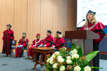 Seven people are participating in an academic ceremony. Six people in red and black gowns and caps are sitting on the stage. One person is standing at a lectern with a microphone and giving a speech. In the background, there is a wooden panel and a screen with text. In the foreground, there are white flowers with green leaves.