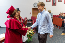 A young man in a light blue shirt shakes hands with a woman in a red gown and biretta. Next to her stands another woman in a black gown and biretta, holding a bag. In the background, there are red chairs, a clock, portraits on the wall, and several people sitting. 