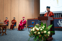 Four people in gowns on the stage of the auditorium. Three are seated. One person—the dean of the faculty—is standing at the lectern and speaking. The university's coat of arms is displayed on the lectern. In the foreground is a large bouquet of white roses and red berries. In the background, part of a screen with the inscription Facultas Pharmaceutica can be seen.