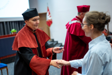 The vice dean shakes hands with a student wearing a blue shirt. The vice dean holds a silver pin in her left hand. Other people can be seen in the background.