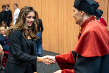 The vice dean shakes hands with a student dressed elegantly in a black jacket. Other people can be seen in the background.