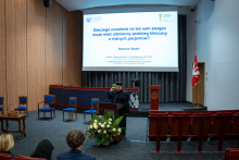 Dr. Białek stands behind the podium on stage. He is wearing a black academic gown. He is giving a speech.  Behind him, a slide with the title of the presentation is displayed on the screen: “Why can an allergy to the same allergen have different clinical manifestations in different patients?”