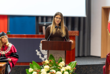 A young, elegantly dressed woman stands at the podium and speaks. A bouquet of flowers can be seen in front of the podium. In the background, there is a multimedia screen and colorful banners.
