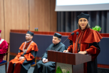The vice dean for teaching in the field of medical analytics, wearing a black and orange gown, stands at the lectern and speaks. Three people in gowns sit in the background.