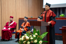 The vice dean for teaching in the field of pharmacy, wearing a black and orange gown, stands at the lectern and gives a speech. Three people in gowns sit in the background.