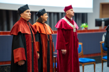 Three people, the vice-rector and vice-deans, dressed in academic gowns, stand in the middle of the auditorium. They are focused and serious. In the background, you can see chairs, a lectern, and a multimedia screen.