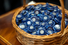 A round wicker basket filled with silver pins. Each pin is packaged in a box with a transparent lid and a navy blue bottom. The pins feature the logo of the Faculty of Pharmacy.