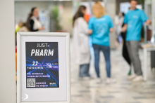 Na zdjęciu widać stojący biały stojak z plakatem promującym wydarzenie „JUST PHARM”, które odbywa się 22 października w holu Wydziału Farmaceutycznego w godzinach 8:00–15:00. Na plakacie znajduje się kod QR i logo Biura Karier. W tle widać kilka osób rozmawiających przy stoisku, część nosi białe fartuchy, a inne niebieskie koszulki. Pomieszczenie jest jasne, z marmurową podłogą i roślinami dekoracyjnymi.