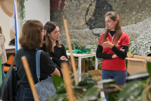 Trzy osoby stojące w pomieszczeniu, rozmawiają. Są nieformalnie ubrane. W tle widać ścianę z mozaiką i kilka stołów.