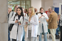 Siedem osób w jasnym holu podczas wydarzenia. Dwie osoby na pierwszym planie noszą białe fartuchy i trzymają ulotki oraz napój. W tle stoją osoby w jasnych i ciemnych ubraniach. Na stołach widoczne są zielone obrusy i materiały informacyjne. W pomieszczeniu znajdują się banery.