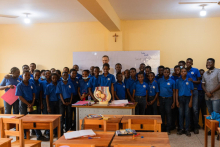 Około 30 osób w sali lekcyjnej pozuje do zdjęcia.Wszyscy się uśmiechają. Większość ubrana jest w niebieskie koszulki. Przed nimi stoją drewniane ławki, na których leżą przybory lekcyjne. Za nimi widać tablicę lekcyjną.