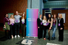 Na zdjęciu znajduje się grupa dziewięciu osób stojących w holu przy ceglanej ścianie. W centrum ustawiony jest pionowy baner w kolorach niebiesko-różowych z napisem „euni well – European University for Well-Being”