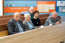 Kilka osób elegancko ubranych siedzi w sali wykładowej. W tle znajduje się napis „dorobek naukowy”.
