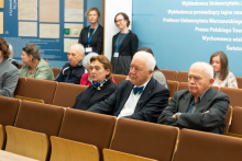 Grupa osób siedzi w audytorium. W pierwszym rzędzie znajdują się trzy starsze osoby – dwóch mężczyzn i kobieta – które uważnie słuchają. 