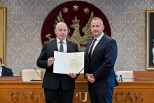 Dwaj mężczyźni stoją obok siebie w formalnym otoczeniu. Osoba po lewej stronie trzyma otwarty dokument skierowany w stronę aparatu.