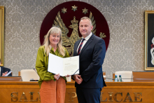 Two people, a woman and a man, are standing next to each other in a formal setting. The person on the left is holding an open document facing the camera.