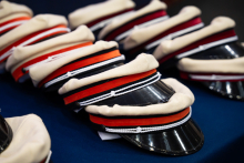 Rows of cream-colored caps with black visors lie on a dark blue table. The caps have bands in red, orange, black, and burgundy. 