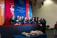 Eleven people—men and women—stand on stage and sing from open sheet music. In front of them, a conductor in a dark suit leads the choir. Colorful banners with the name of the university and the inauguration slogan hang in the background. Academic caps lie on a table in front of the stage. The audience sits on the right.