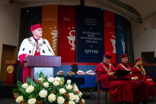 The rector, wearing a red biretta and a red coat with a white collar, speaks at the podium. Behind him sit three people in red gowns and birettas. In the background, there are colorful banners with the names of the departments and the inscription “Inauguration of the Academic Year 2025/2026.” In front of the podium, there is a bouquet of white roses with red accents.