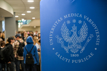 In the foreground, there is a large blue banner with a white WUM logo and the inscription “Universitas Medica Varsoviensis Saluti Publicae 1809.” In the background, there are several people in the hallway, some of them talking at the stands.