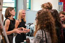 Several people are standing around a table in a bright room. Two people in black dresses are gesturing and explaining something to the group. There are small objects and a brown bottle on the table.